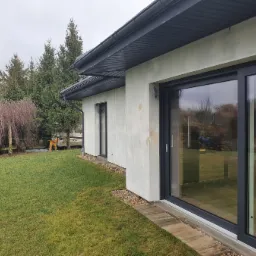 Nowoczesny dom jednorodzinny z dużymi, przesuwnymi oknami w grafitowych ramach, widok z zewnątrz na trawnik i drzewa w tle, elewacja w trakcie wykańczania.