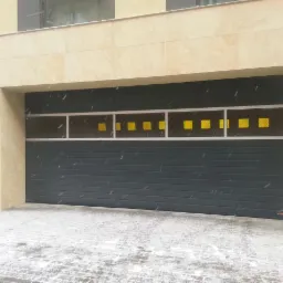 Zimowy widok na nowoczesną, szarą bramę garażową z poziomymi panelami i kwadratowymi, żółtymi przeszkleniami, z delikatnym opadem śniegu.