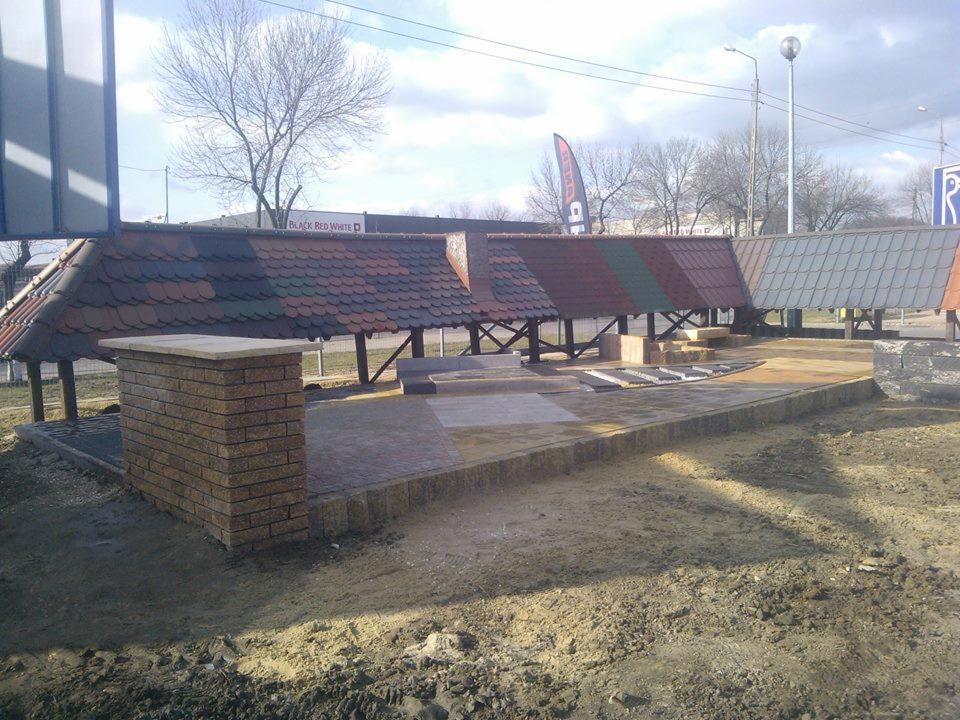 Ekspozycja altan z różnymi pokryciami dachowymi i kostki brukowej na placu. Widoczne różne kolory i wzory materiałów wykończeniowych.