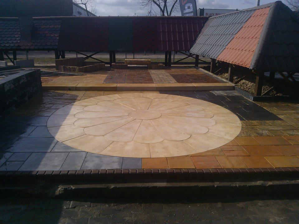 Wzorzysta, okrągła kostka brukowa w kształcie kwiatu na patio, otoczona różnymi kolorami i wzorami kostki. Prezentacja możliwości aranżacji nawierzchni.