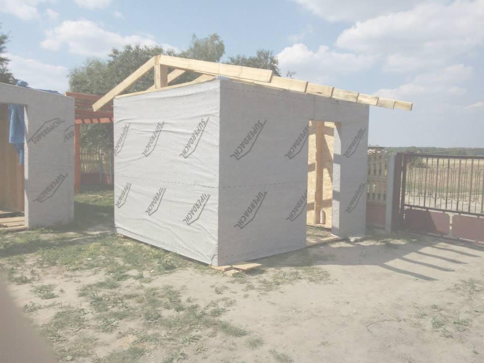 Konstrukcja małego garażu blaszanego w trakcie budowy, z widoczną drewnianą ramą i membraną Superdachl na ścianach. W tle ogrodzenie i zieleń.