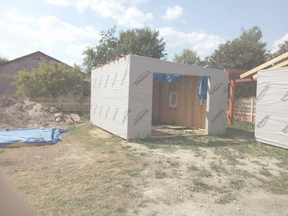 Budowa garażu blaszanego w trakcie: konstrukcja szkieletowa obłożona membraną, widoczny fragment dachu i okno, w tle dom i drzewa.