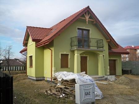 Jasnozielony dom jednorodzinny z czerwonym dachem i balkonem, widoczny stos drewna i skrzynka gazowa na niezagospodarowanej działce.