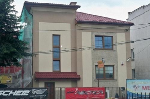 Dwupiętrowy dom z beżową elewacją, częściowo zakryty siatką ochronną podczas prac, widoczne banery reklamowe w tle.