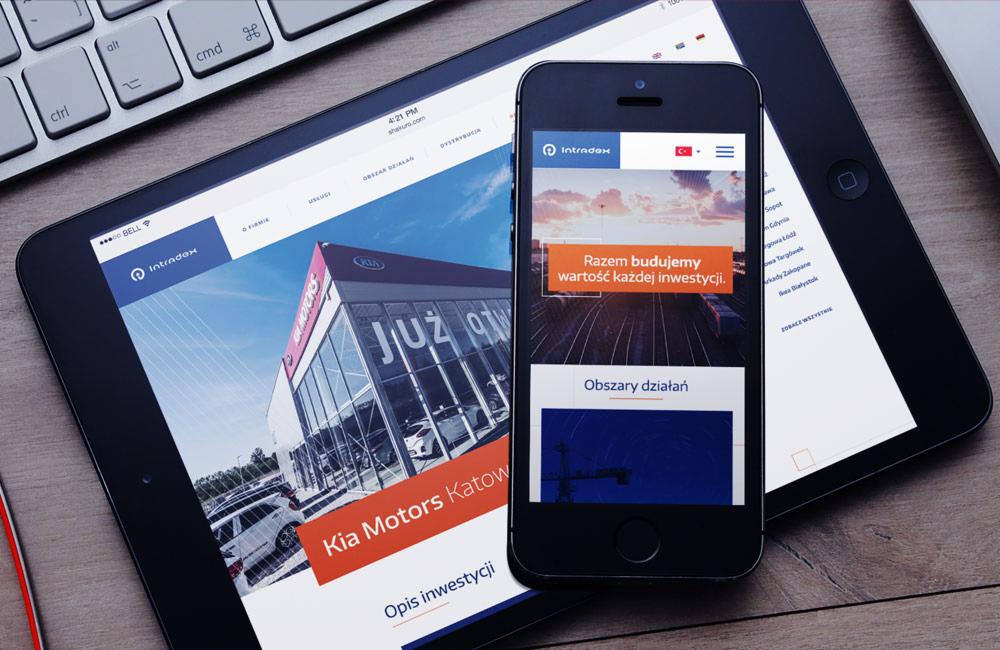 Tablet i smartfon wyświetlają responsywną witrynę Intradex z widokiem na salon Kia Motors Katowice, leżące obok klawiatury na drewnianym biurku.