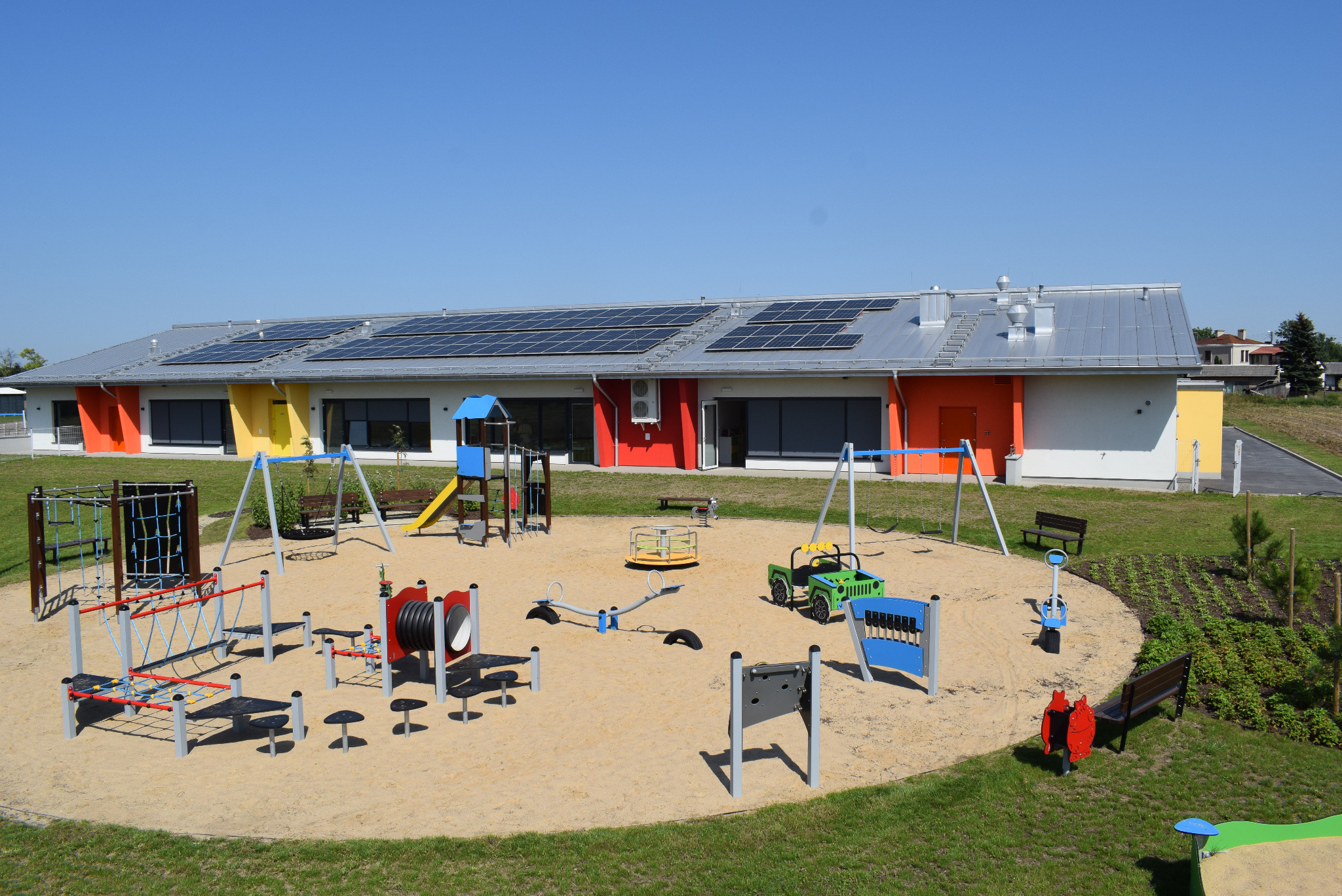 Nowoczesny budynek przedszkola z panelami słonecznymi na dachu, kolorową elewacją i placem zabaw z piaskiem oraz różnorodnymi urządzeniami do zabawy.