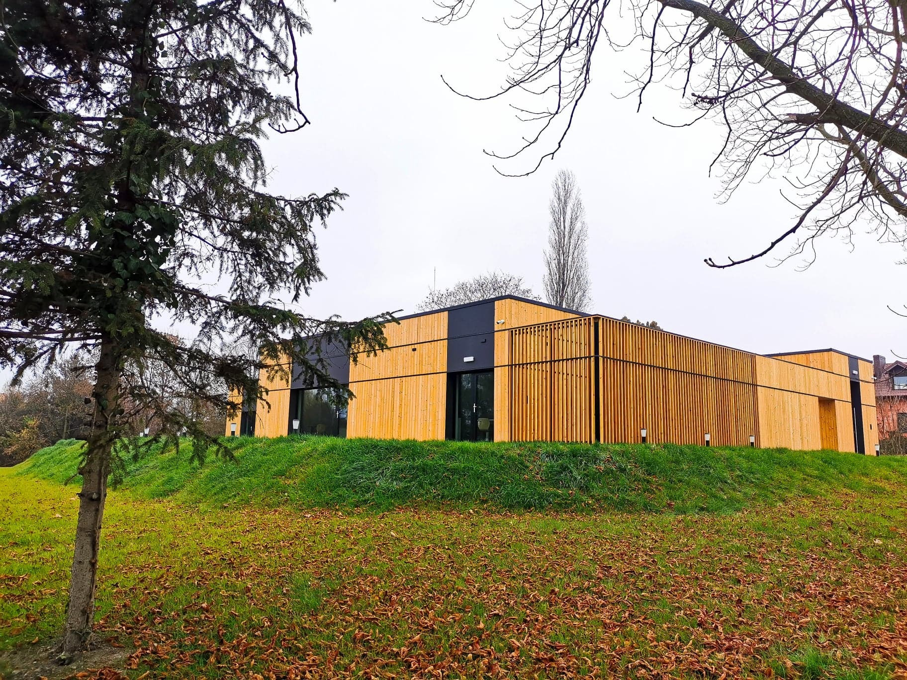 Nowoczesny budynek z drewnianą elewacją i czarnymi akcentami, otoczony zielenią i drzewami, sfotografowany w pochmurny dzień. Widoczny fragment trawnika z opadłymi liśćmi.