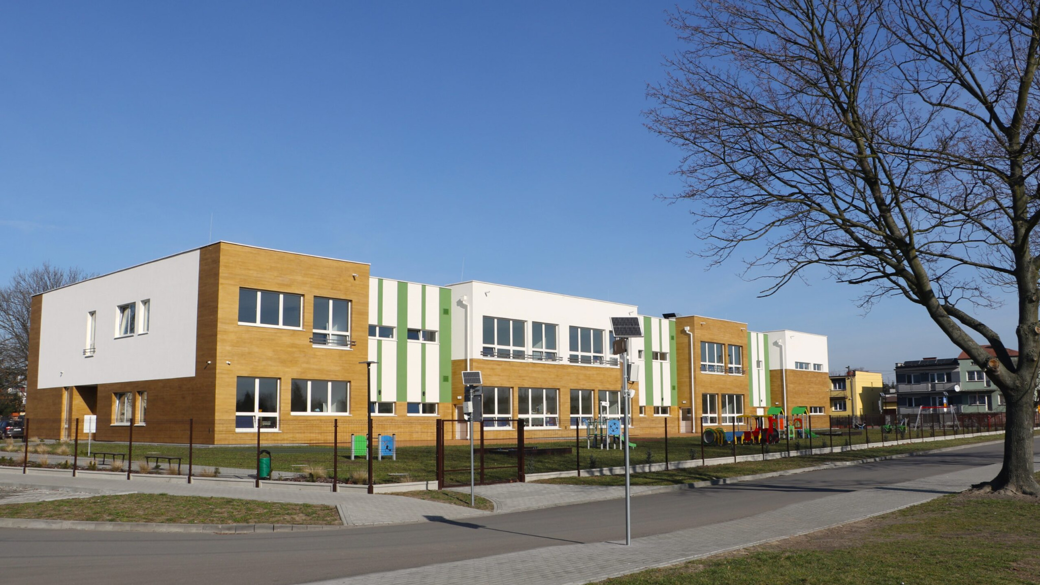 Nowoczesny budynek przedszkola integracyjnego z elementami drewna i zieleni, widok z zewnątrz w słoneczny dzień. Na dachu panele słoneczne, plac zabaw w tle.