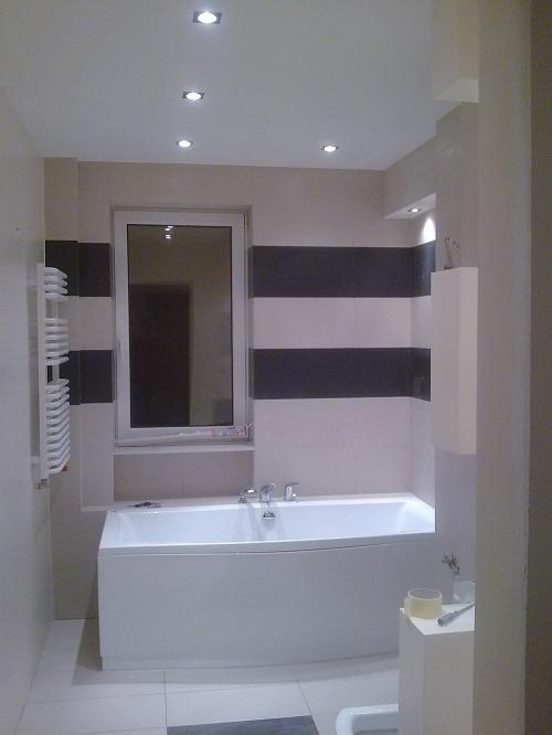 Nowoczesna łazienka z białą wanną, panelami w odcieniach szarości i białymi płytkami podłogowymi, oświetlona punktowymi lampami sufitowymi.