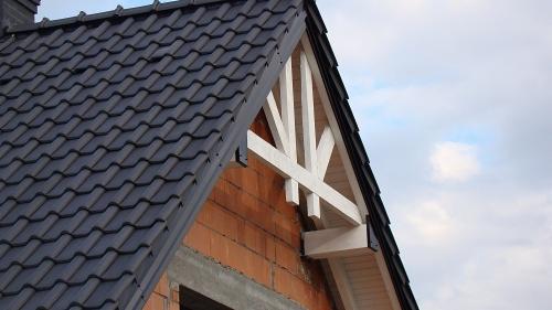 Częściowy widok dachu z czarną dachówką ceramiczną, białymi elementami więźby dachowej i fragmentem muru z czerwonej cegły w trakcie budowy, na tle pochmurnego nieba.