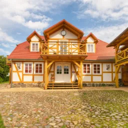 Elewacja domu w stylu rustykalnym z czerwonym dachem i drewnianymi elementami, widok z brukowanego podjazdu w słoneczny dzień. Na ścianie dekoracyjny zegar.