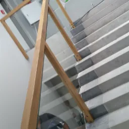 Szklana balustrada z drewnianymi elementami mocującymi na schodach wyłożonych szaro-białą mozaiką, widok z dołu.