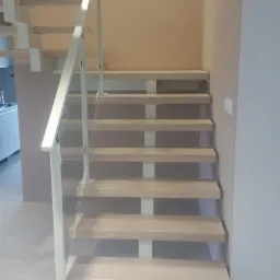 Nowoczesne schody wewnętrzne z drewnianymi stopniami i białą metalową balustradą, widok z dołu na jasnym tle ściany.