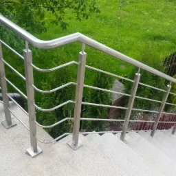 Zewnętrzna balustrada schodowa ze stali nierdzewnej z poziomymi prętami o falistym kształcie, zamontowana na granitowych stopniach, z widokiem na zielony trawnik i tuję.