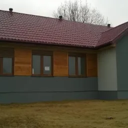 Niski, parterowy dom z drewnianą elewacją, kryty czerwoną blachodachówką. Widoczne okna z roletami i wejście z drewnianymi schodkami. Tło stanowi niebo i drzewa.