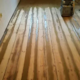 Drewniana podłoga w trakcie renowacji z widocznymi śladami po szlifowaniu i maszyną do szlifowania parkietu w prawym górnym rogu.