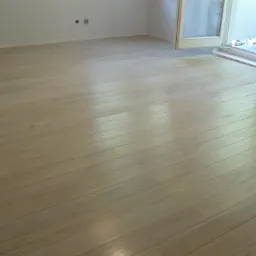 Jasna, drewniana podłoga z widocznymi słojami, ułożona w pokoju z białymi ścianami i otwartym balkonem, widoczne dwa otwory na gniazdka elektryczne na ścianie.