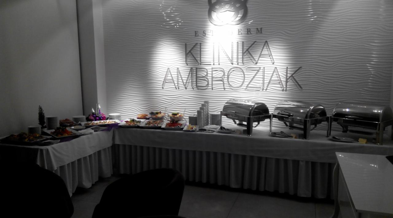 Stół cateringowy z przekąskami i podgrzewaczami potraw w sali z dekoracyjną ścianą i logo 'Klinika Ambroziak'.
