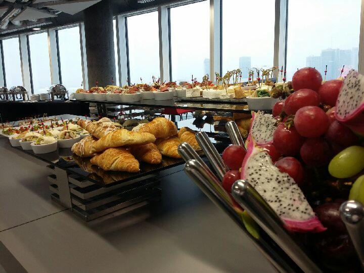 Bogato zastawiony stół z przekąskami, croissantami i owocami egzotycznymi na tle okien z widokiem na miasto.