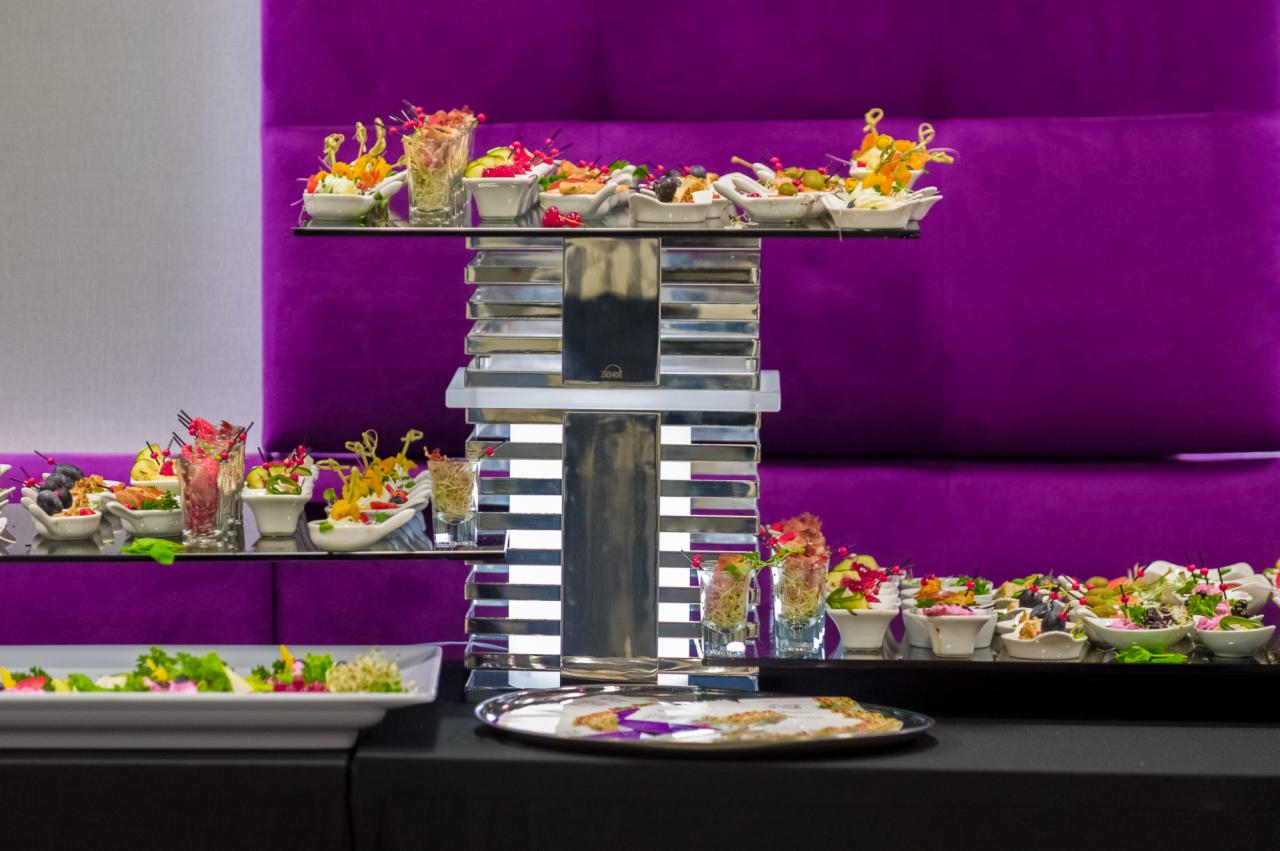 Elegancki bufet z przekąskami na srebrnym, kaskadowym stojaku, z małymi porcjami sałatek i kanapek na białych naczyniach, ustawiony na czarnym stole przed fioletową tapicerowaną ścianą.
