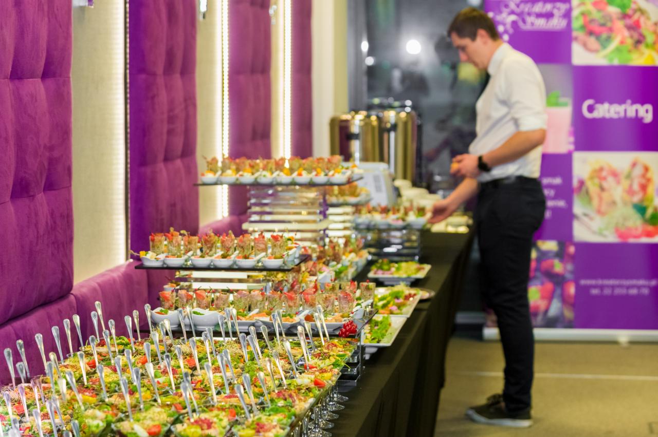 Stół cateringowy z różnorodnymi przekąskami, w tle osoba w białej koszuli i czarnych spodniach, fioletowa ściana dekoracyjna i banner z napisem Catering.