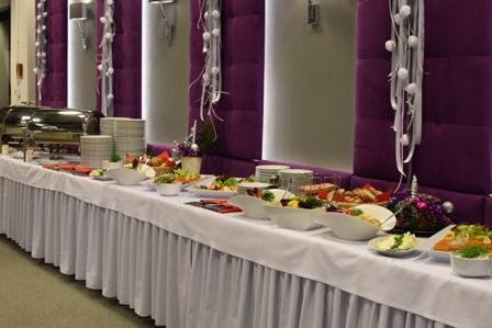 Długi stół cateringowy z białym obrusem, zastawiony różnorodnymi potrawami w miseczkach i na talerzach, w tle fioletowe panele dekoracyjne i srebrne kolumny z ozdobami.