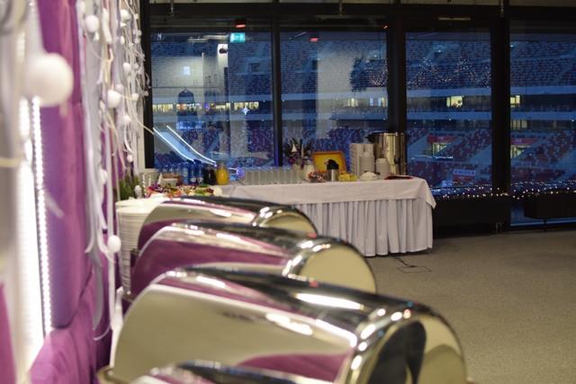 Bufet z podgrzewaczami potraw i napojami na tle stadionu oświetlonego nocą, dekoracje w kolorze fioletowym.