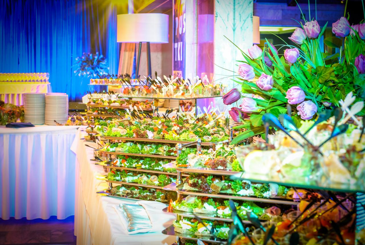 Bogato zastawiony stół cateringowy z wielopoziomowymi paterami, na których znajdują się różnorodne przekąski, sałatki i finger food, udekorowany bukietem różowych tulipanów, oświetlony niebieskim...