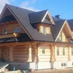 Dwukondygnacyjny dom z bali w stylu góralskim z ciemnym dachem, widoczne schody wejściowe i fragment terenu w trakcie zagospodarowywania.