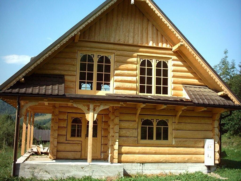 Gotowy, dwukondygnacyjny dom z bali z brązowym dachem, widoczne charakterystyczne okna z łukami na górze i podcień z drewnianymi kolumnami, w tle zalesione wzgórza.