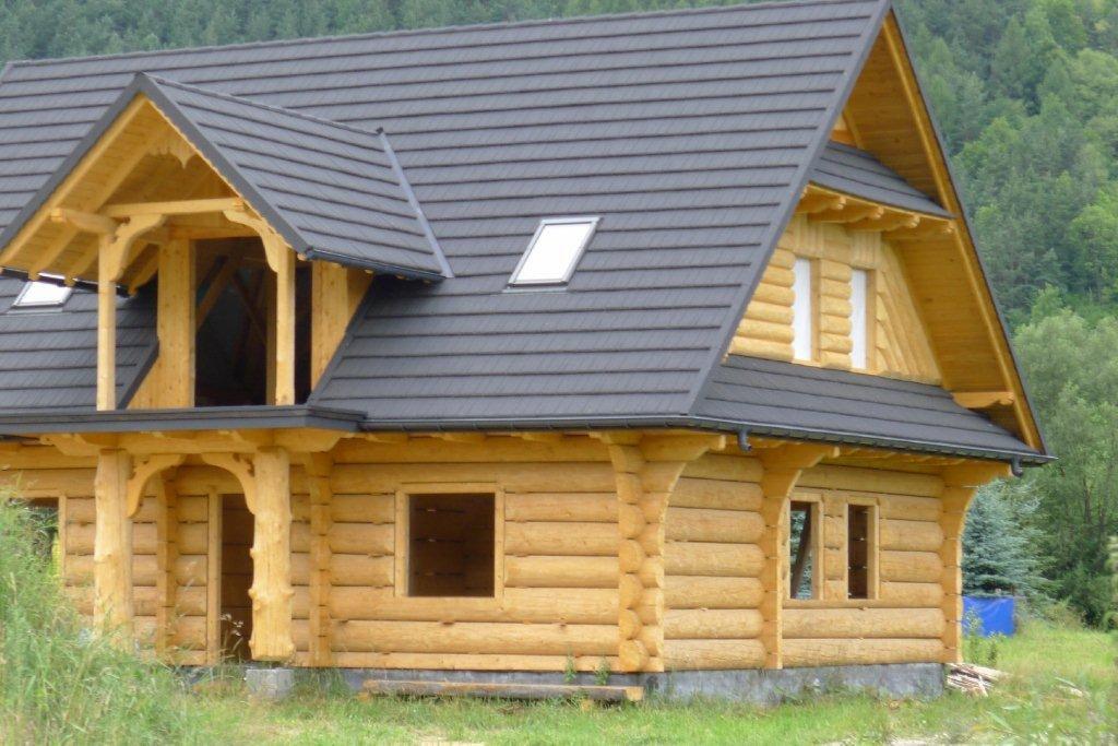 Surowy stan budowy domu z bali z ciemnoszarą, metalową dachówką, widoczne drewniane elementy konstrukcyjne i okno dachowe, tło stanowi zieleń drzew.