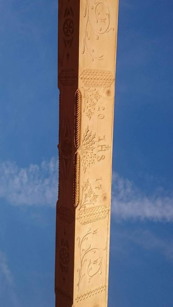 Drewniany słup z rzeźbieniami roślinnymi i geometrycznymi na tle błękitnego nieba.