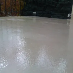 Jasna, gładka powierzchnia balkonu po renowacji z widocznymi refleksami światła. W tle drzewa i fragment budynku.