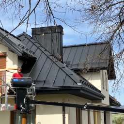 Usługi Blacharsko-Dekarskie - Pracownik na podnośniku koszowym przy dachu domu z czarną blachą trapezową i kominem. Widoczne gałęzie drzew i błękitne niebo.