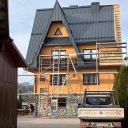 Usługi Blacharsko-Dekarskie - Dom z żółtą elewacją i szarym dachem w trakcie prac dekarskich. Widoczne rusztowania, drabiny i samochód dostawczy. Część dachu pokryta drewnianymi łatami.