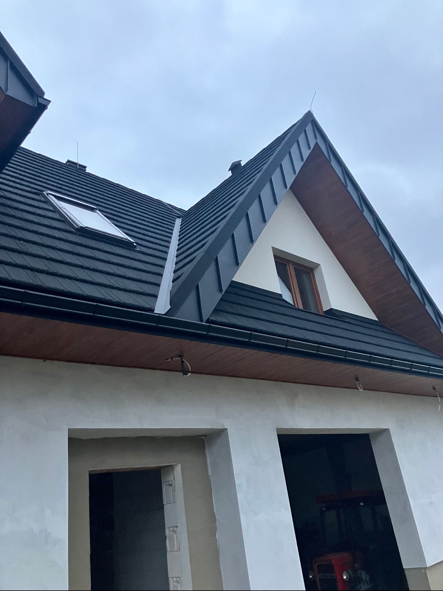 Nowy dach z czarnej dachówki ceramicznej, okno dachowe i wykończenia z drewna. Budynek w trakcie budowy, widoczne surowe ściany i otwory okienne. Pochmurne niebo w tle.