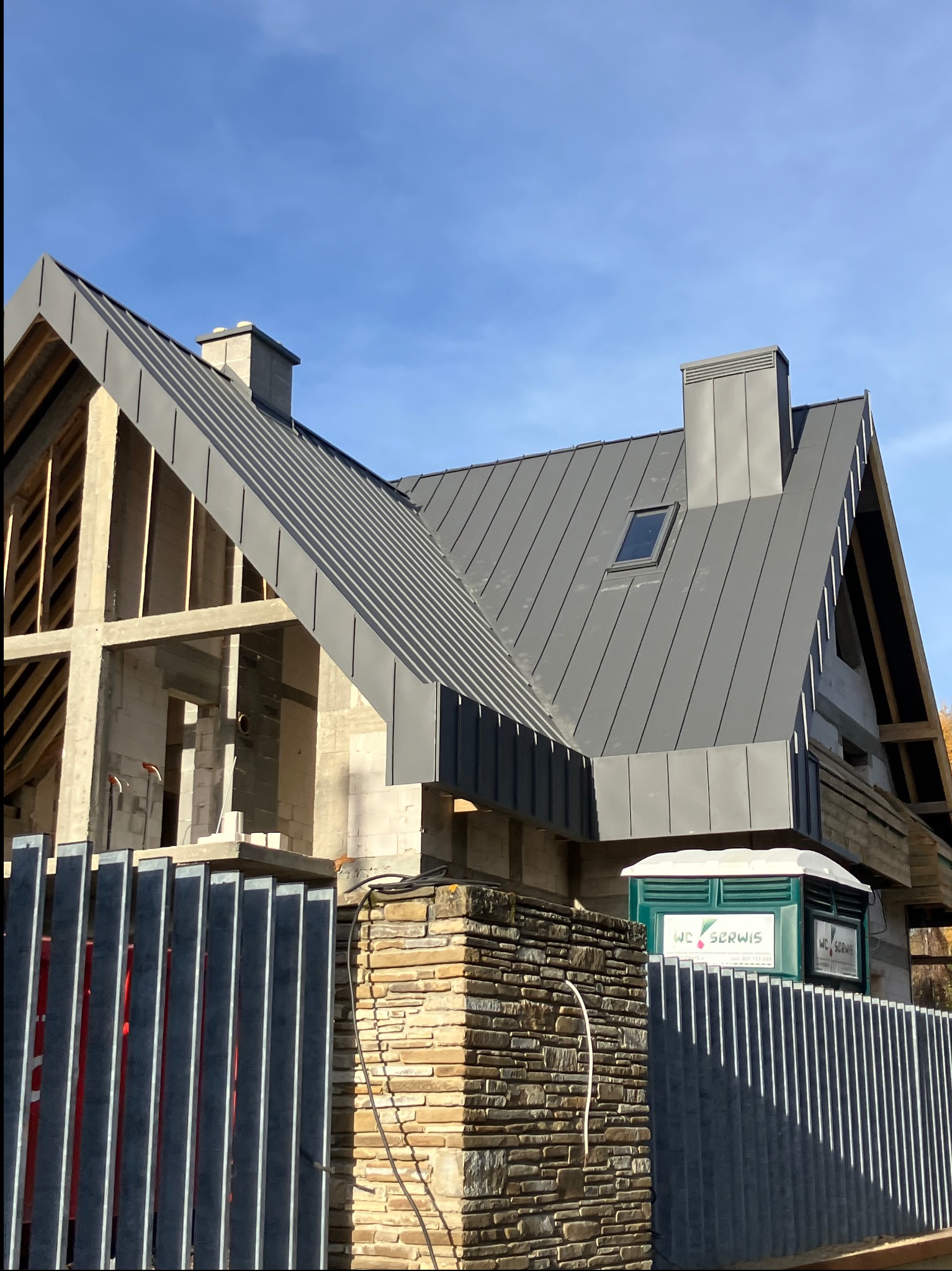 Surowy dom murowany z szarą, metalową konstrukcją dachu i kominem, widoczny przez metalowe ogrodzenie. Na zewnątrz toaleta WC Serwis.