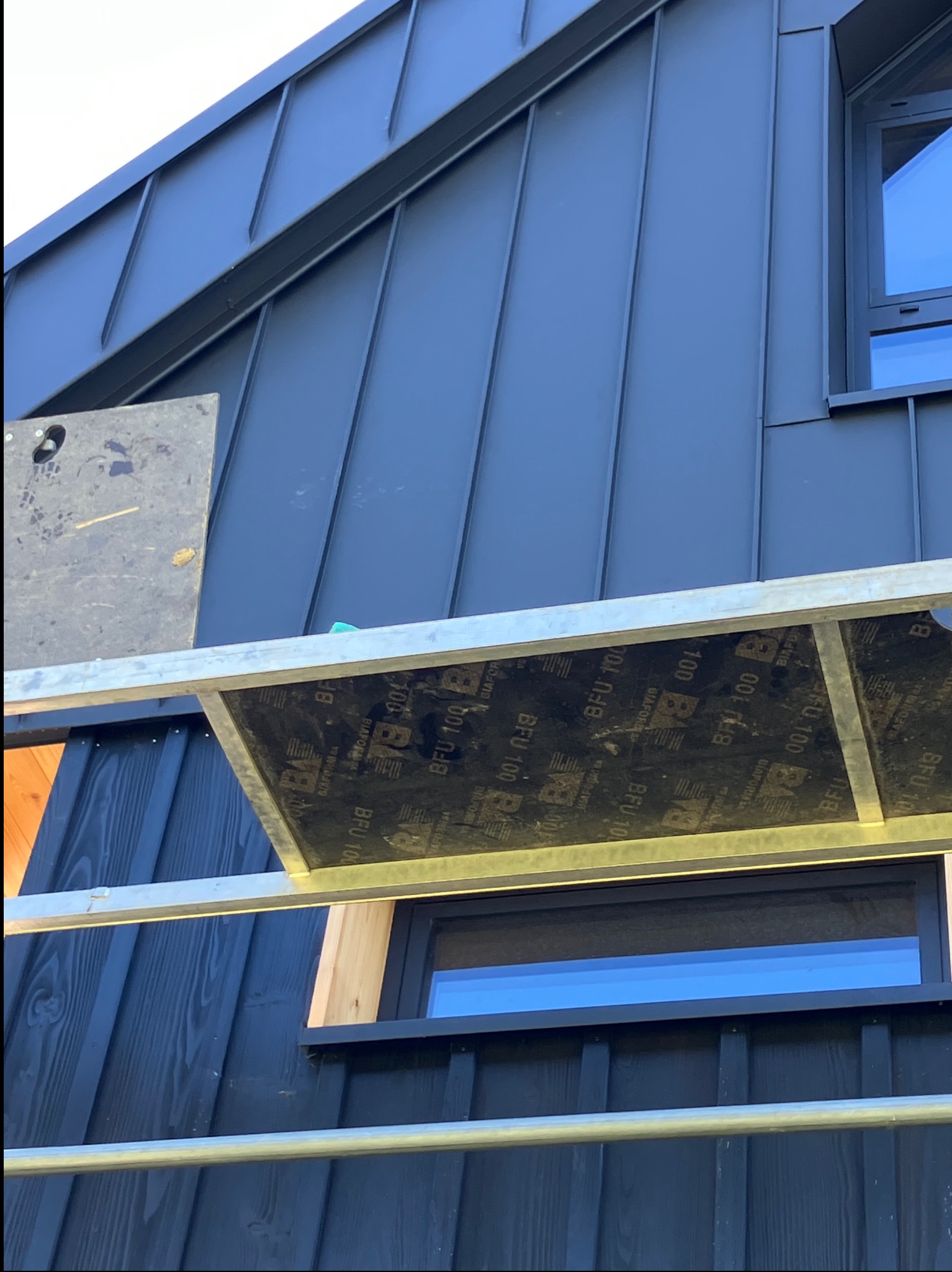 Ciemny, metalowy dach domu z widocznym oknem i fragmentem rusztowania. Detal pokrycia dachowego z blachy na tle drewnianej elewacji.