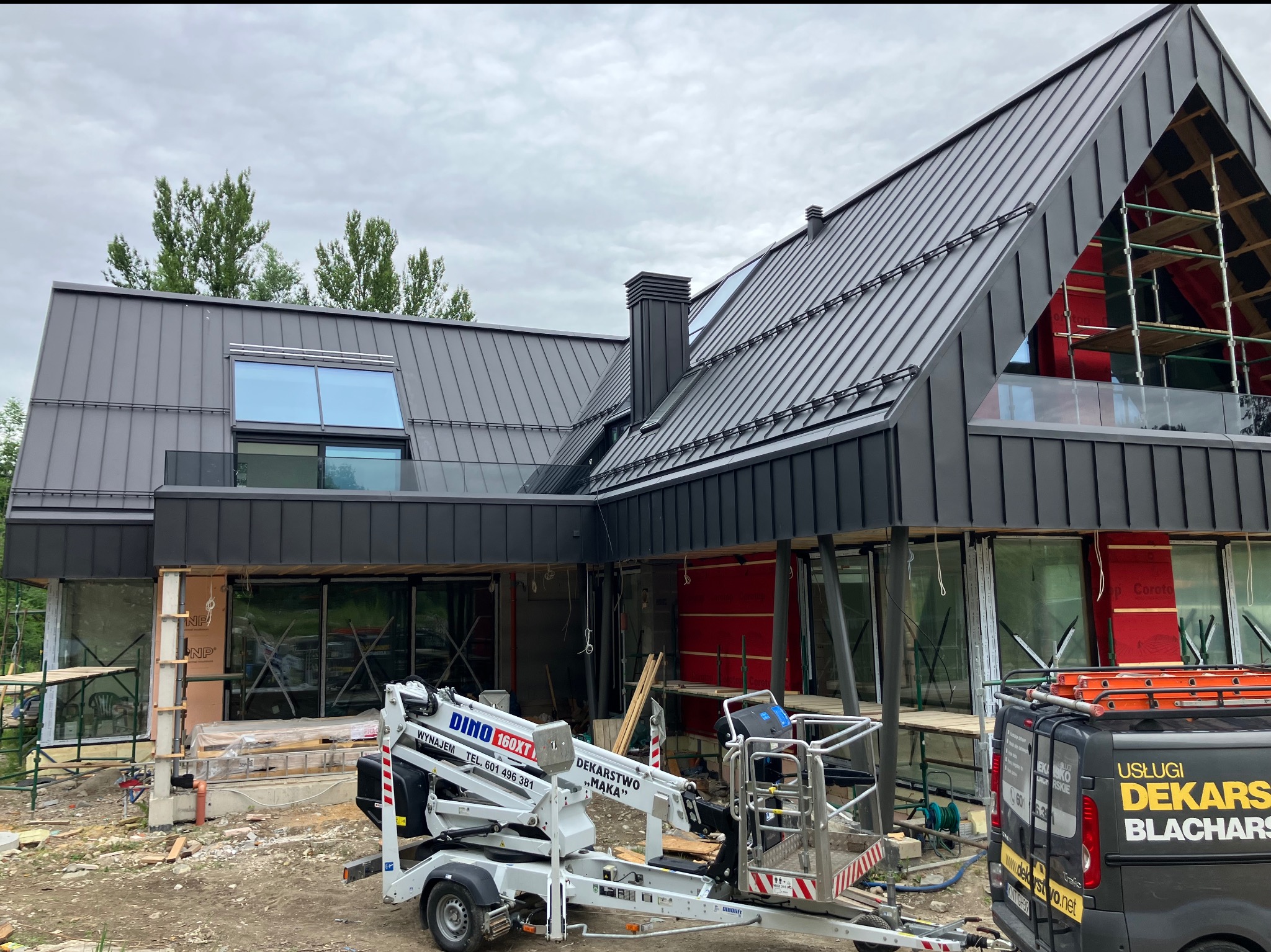 Nowoczesny dom w trakcie budowy z ciemnym dachem z blachy na rąbek stojący. Widoczny podnośnik koszowy i samochód dekarski.