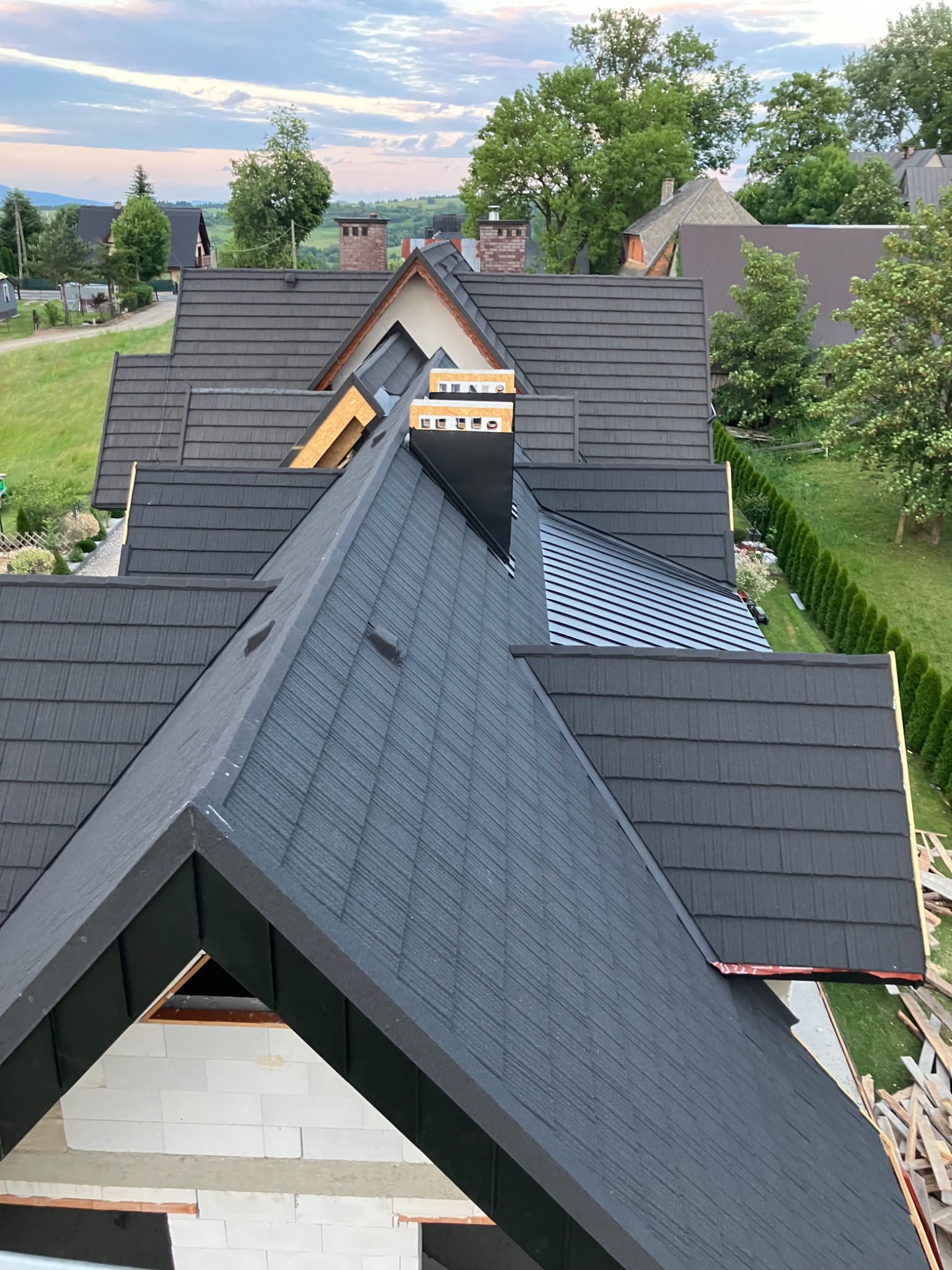 Nowoczesny, grafitowy dach z blachodachówki modułowej, widok z góry na ukończony fragment i surowy stan poddasza. W tle zabudowa jednorodzinna i zieleń.