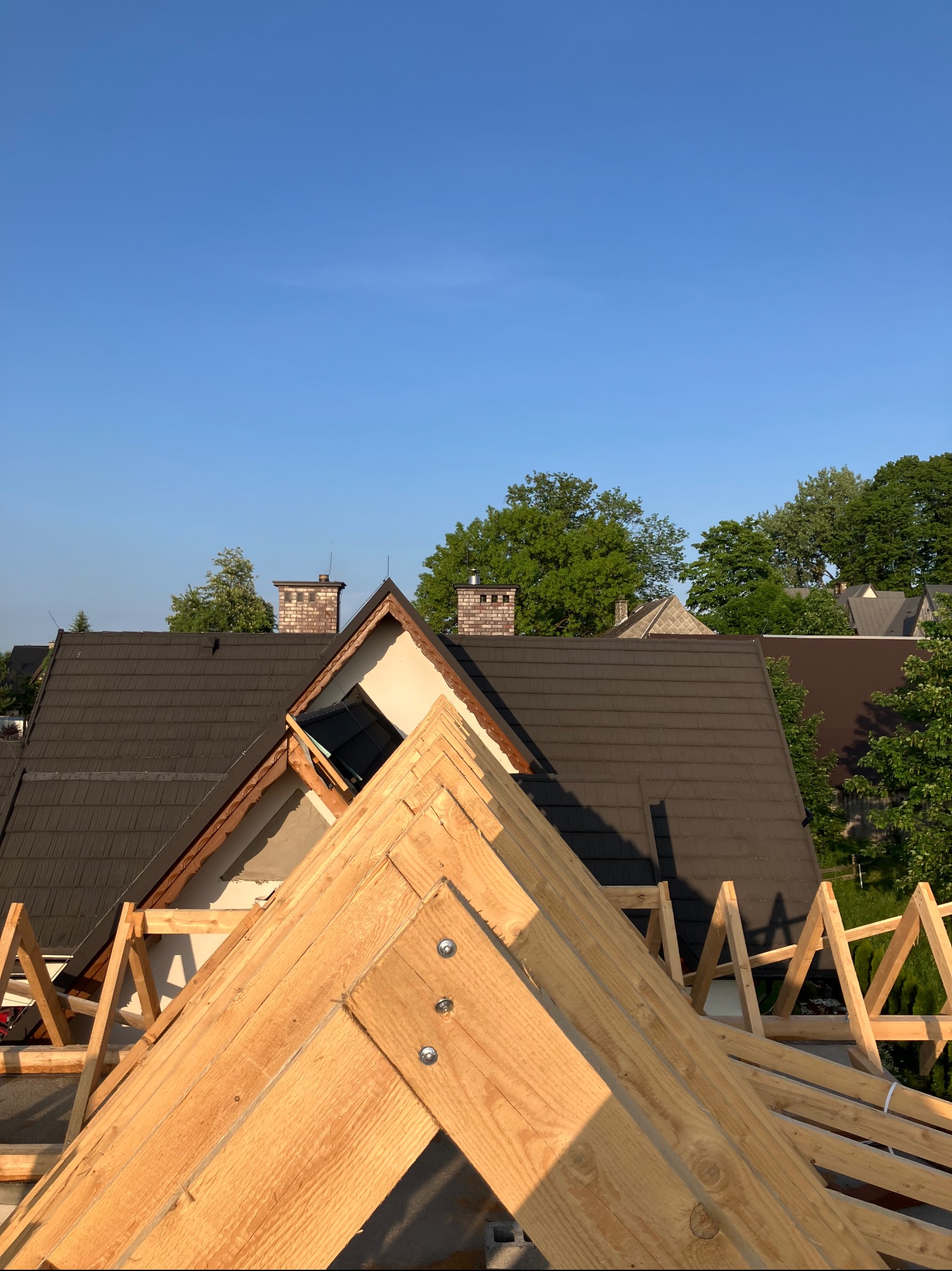 Wiązar dachowy z jasnego drewna na tle ciemnego dachu z dachówki i błękitnego nieba. Widoczne kominy i zieleń drzew w oddali. Konstrukcja w trakcie montażu.