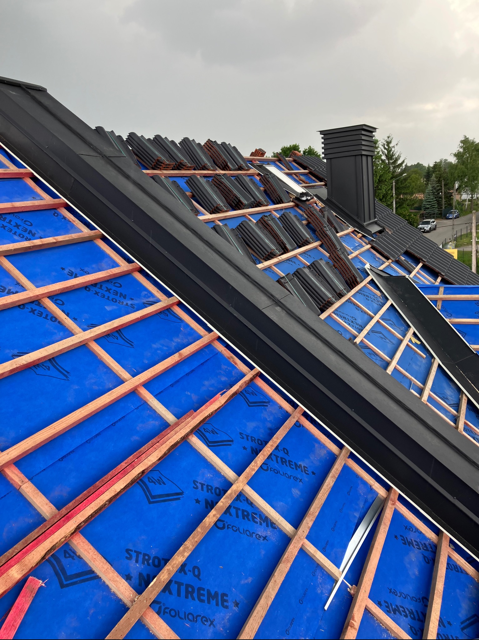 Montaż czarnej dachówki na niebieskiej membranie w Szaflarach. Widoczna drewniana konstrukcja i komin. Częściowo pokryty dach w trakcie prac dekarskich.