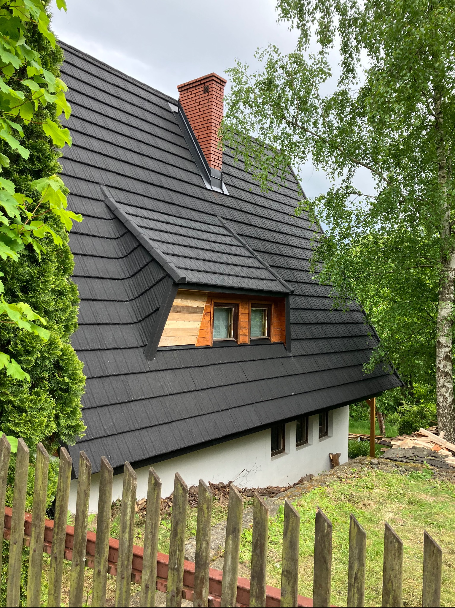 Czarny dach z drewnianymi oknami na poddaszu, ceglany komin, widok zza drewnianego płotu i zieleni. Część dachu poza kadrem, co dodaje unikalności ujęciu.