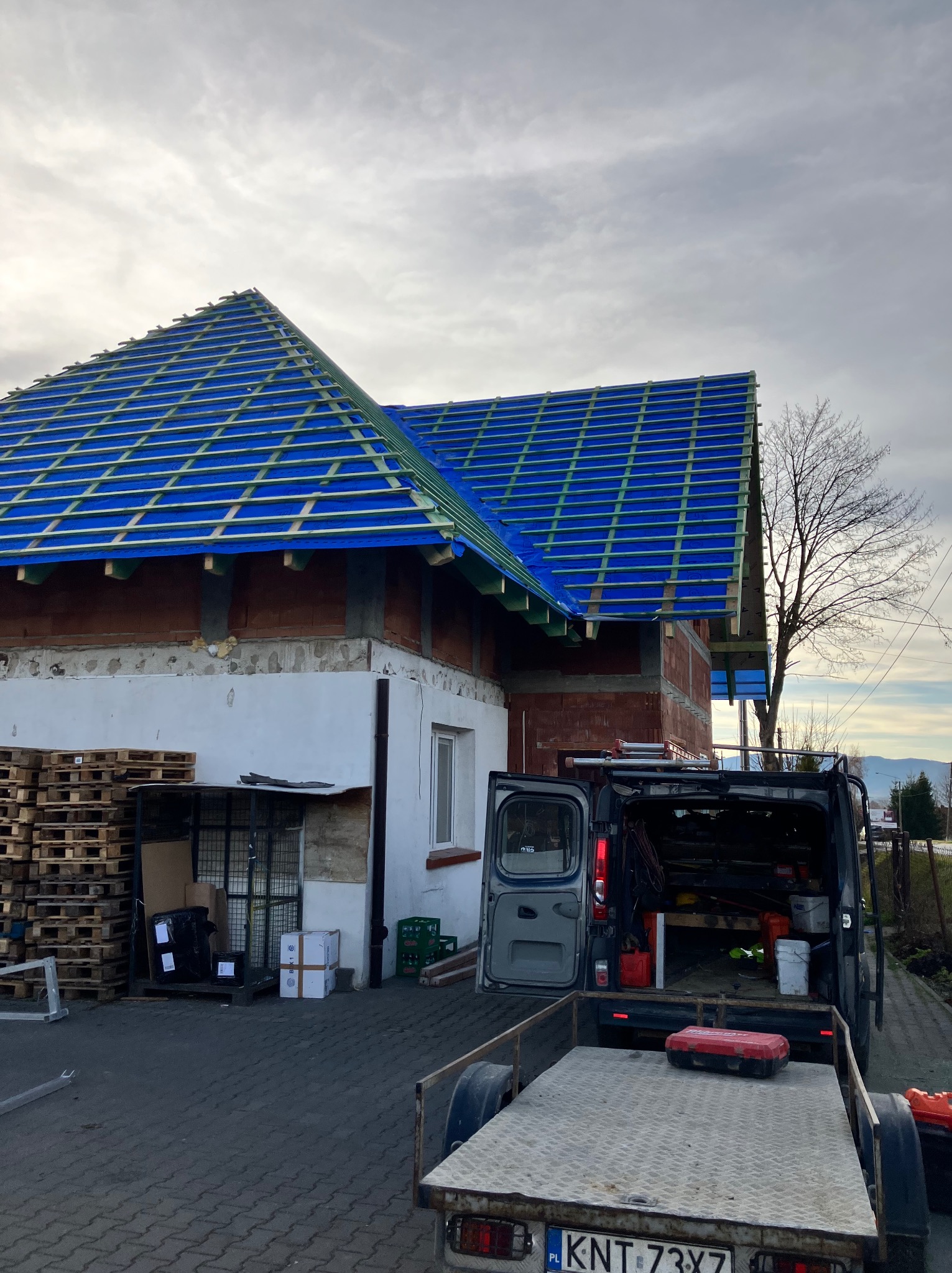 Dom w trakcie krycia dachu, widoczna niebieska membrana i łaty. Na pierwszym planie przyczepka i samochód dostawczy z narzędziami. Palety z materiałami budowlanymi obok budynku.