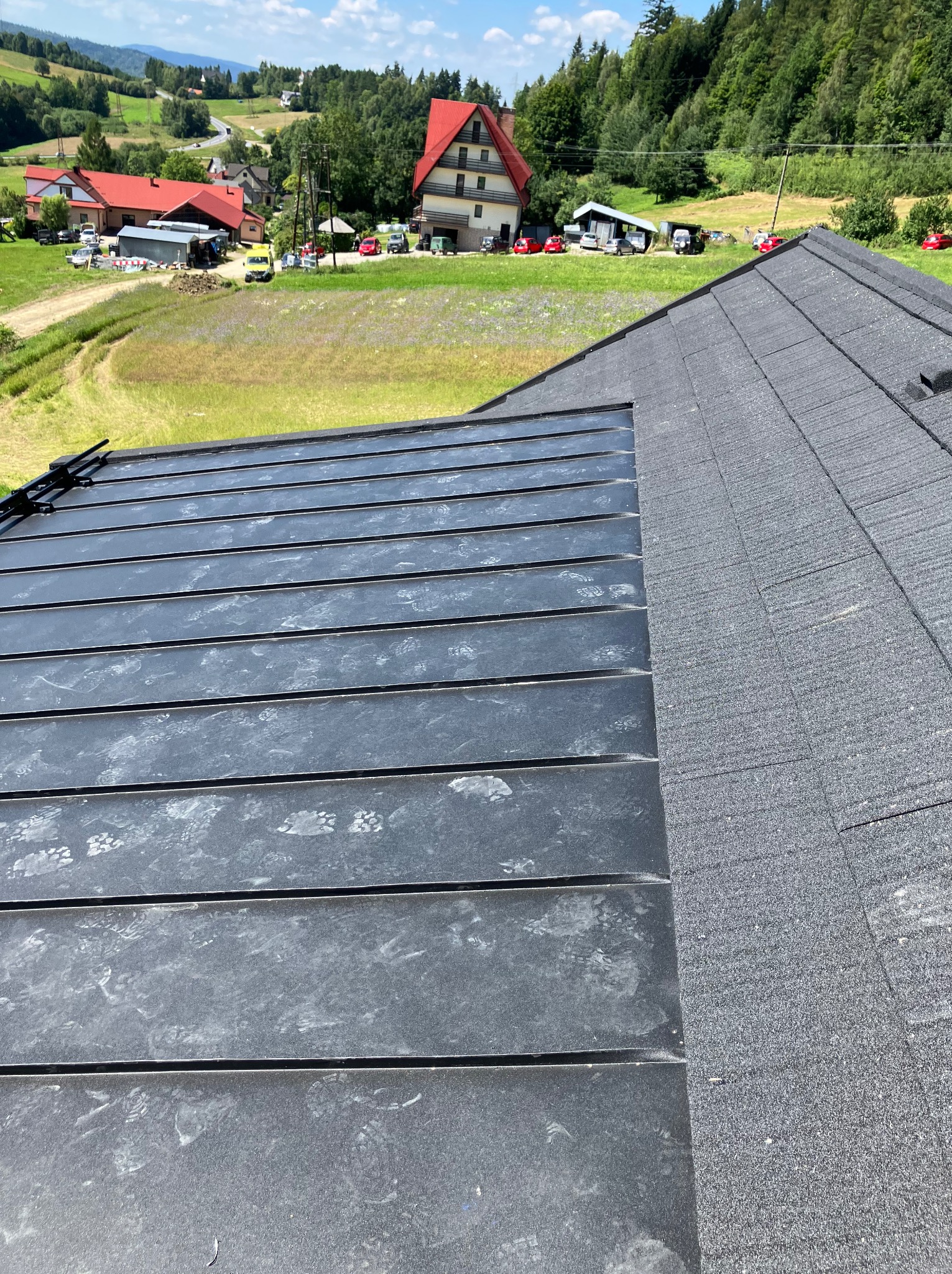 Fragment dachu z blachy na rąbek stojący i dachówki bitumicznej na tle krajobrazu wiejskiego z domami i zielenią. Widoczne ślady montażu na blasze.