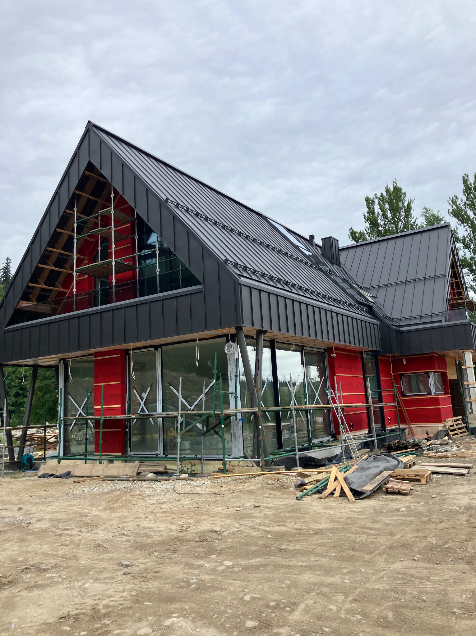 Nowoczesny dom w trakcie budowy z czarnym dachem z blachy na rąbek stojący. Widoczne rusztowania i elementy konstrukcyjne dachu. Czerwone płyty na ścianach.