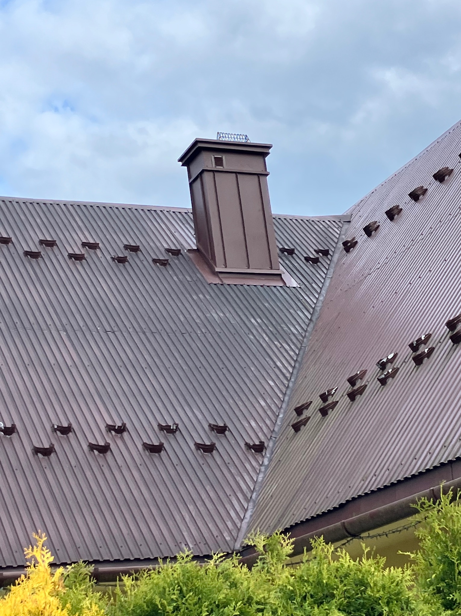 Brązowy, blaszany dach z kominem i zabezpieczeniami przeciwśniegowymi, widoczny z dołu na tle pochmurnego nieba i zieleni.