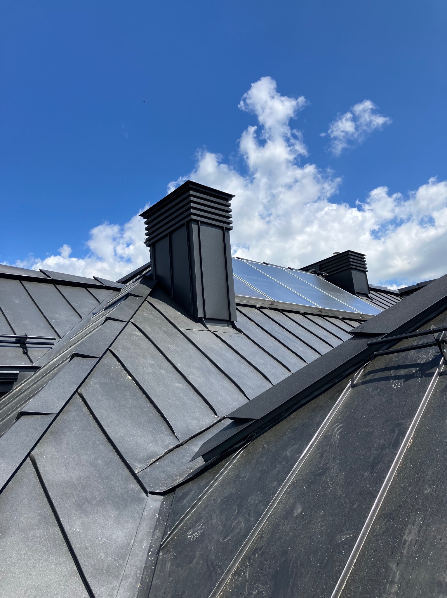 Szczegółowe ujęcie czarnego, metalowego dachu z kominami na tle błękitnego nieba z chmurami. Widoczne panele słoneczne. Dach pokryty blachą na rąbek stojący.