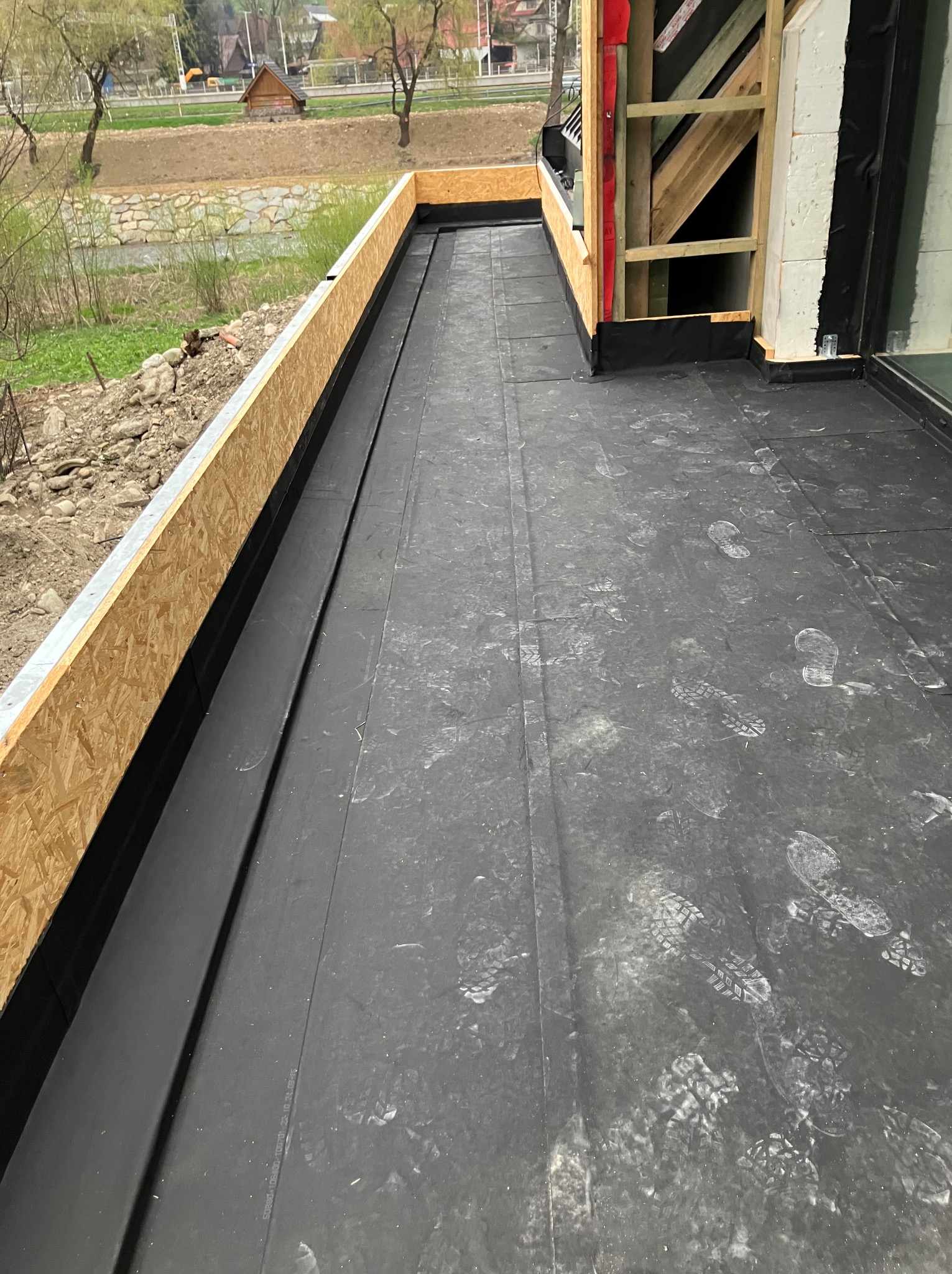 Czarna membrana dachowa z widocznymi śladami butów, obramowana płytą OSB. W tle widoczny krajobraz z rzeką i zabudowaniami.