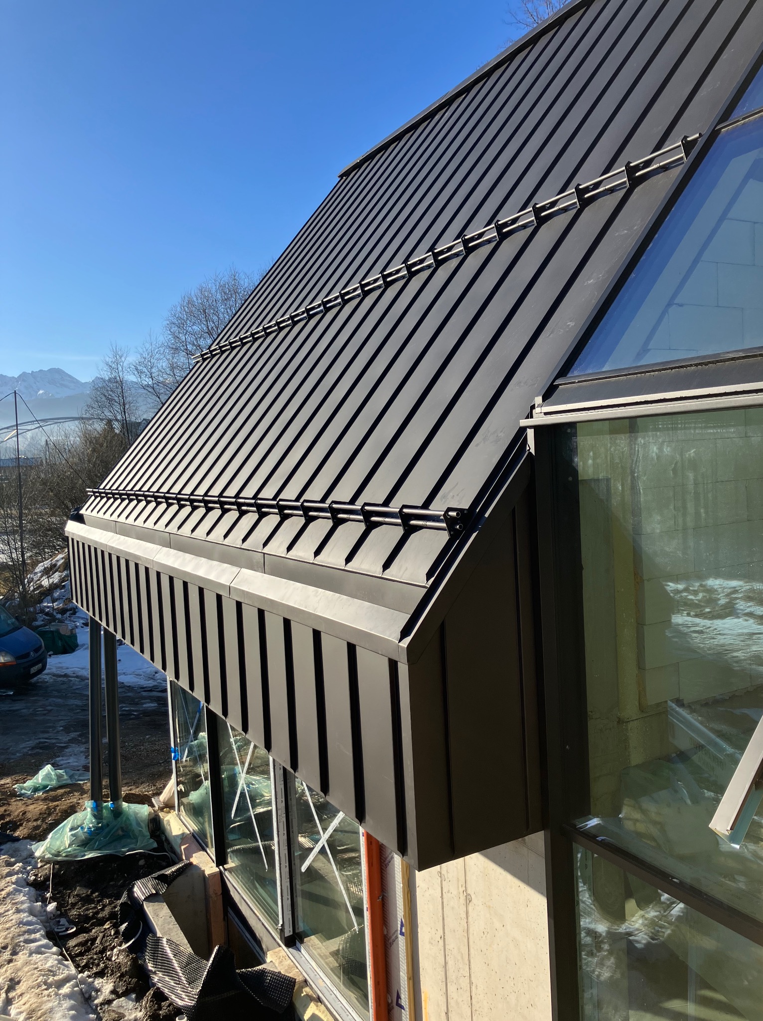 Czarny, metalowy dach z widocznymi mocowaniami na tle błękitnego nieba. Nowoczesna architektura z dużymi oknami i elementami wykończeniowymi. Widok na góry w oddali.